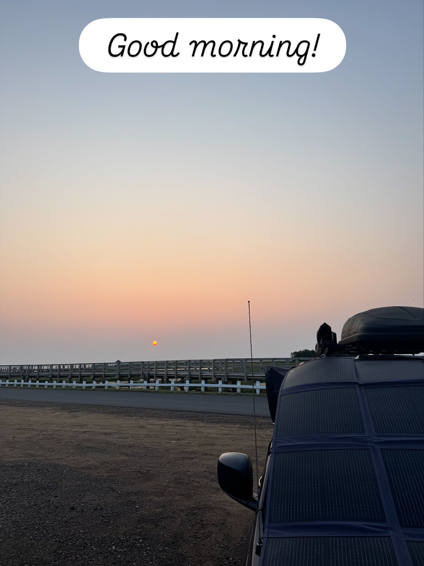 Blue Meanie van parked at sunrise on a beach with bridge behind it.