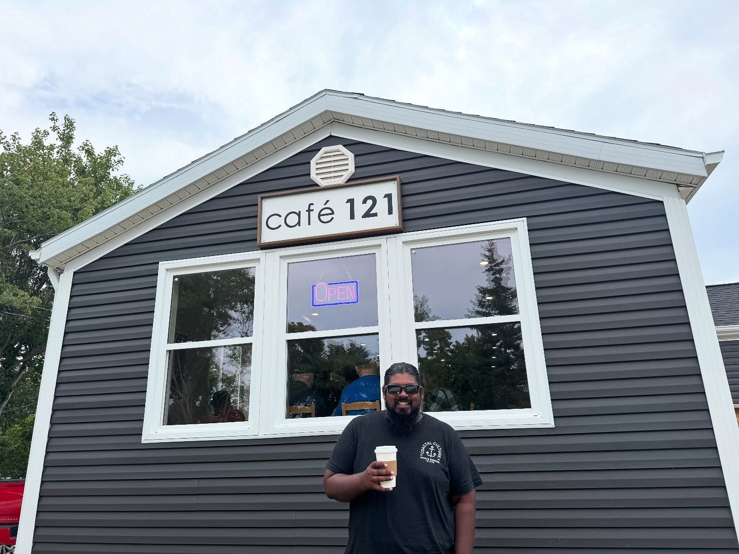 Man in black shirt holds coffee cup in front of Café 121 storefront.