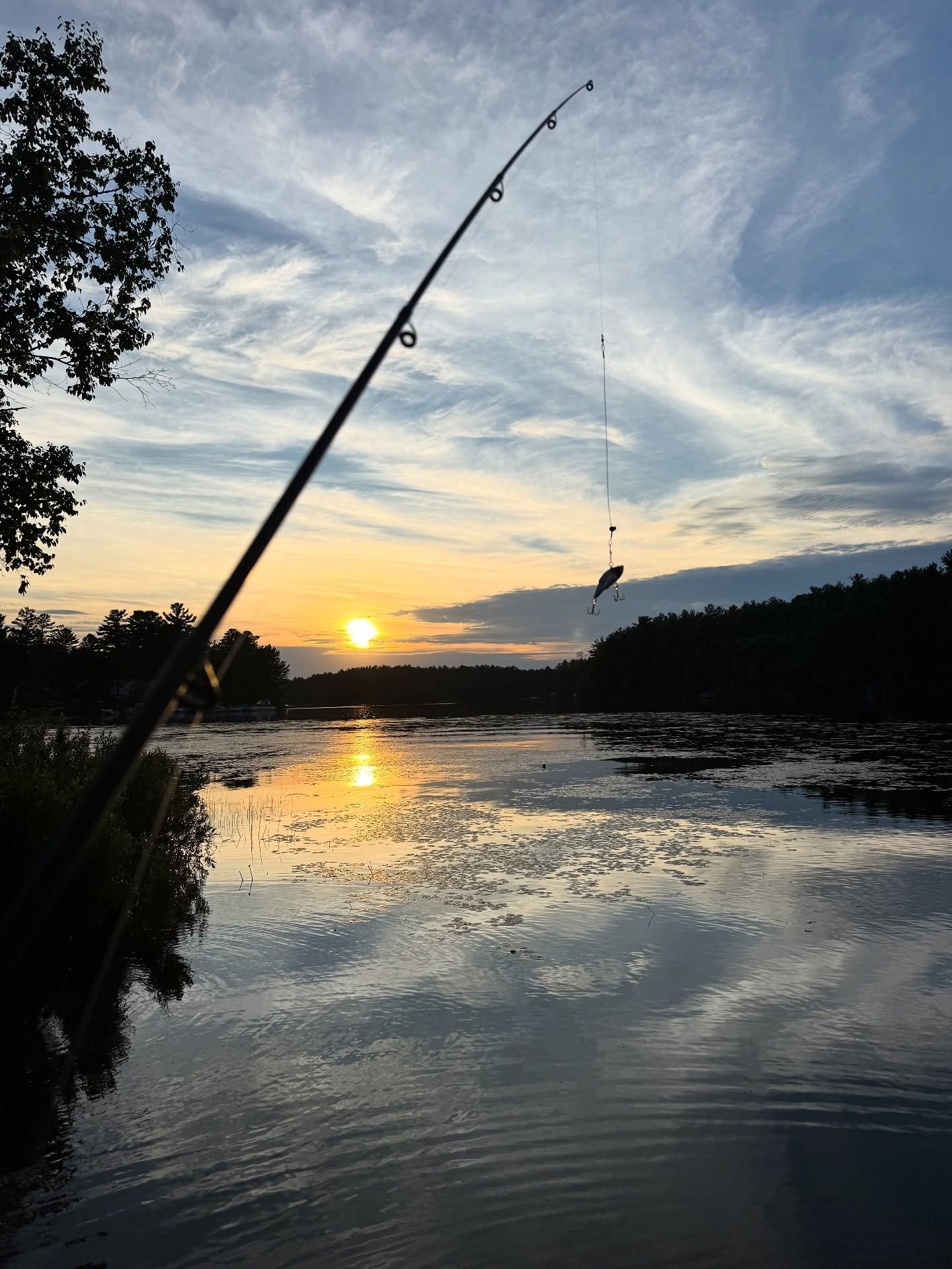 A fishing rod extends over a calm river at sunset with golden sky and treeline.