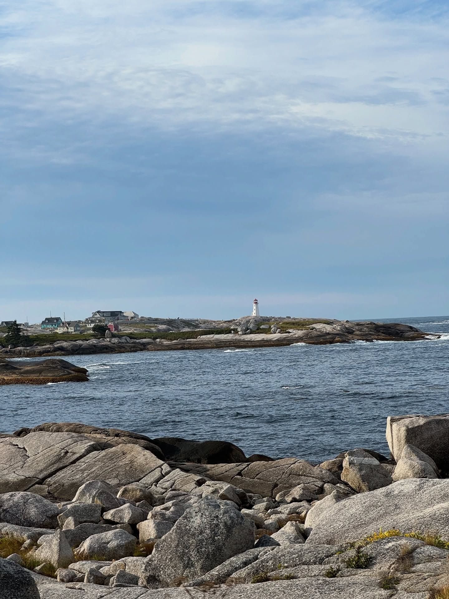Rocky coastline with lighthouse, houses, and calm blue-green water under cloudy sky.