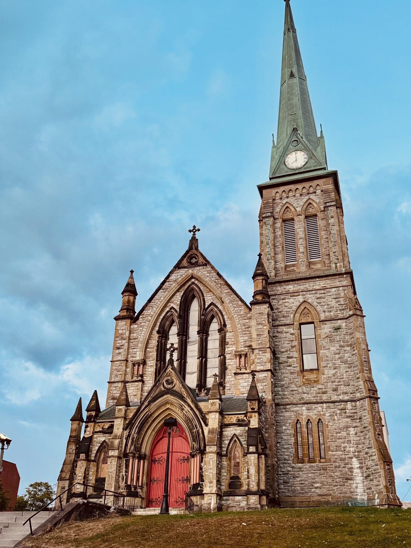 Stone church building with tall spire and red entrance doors against blue sky.