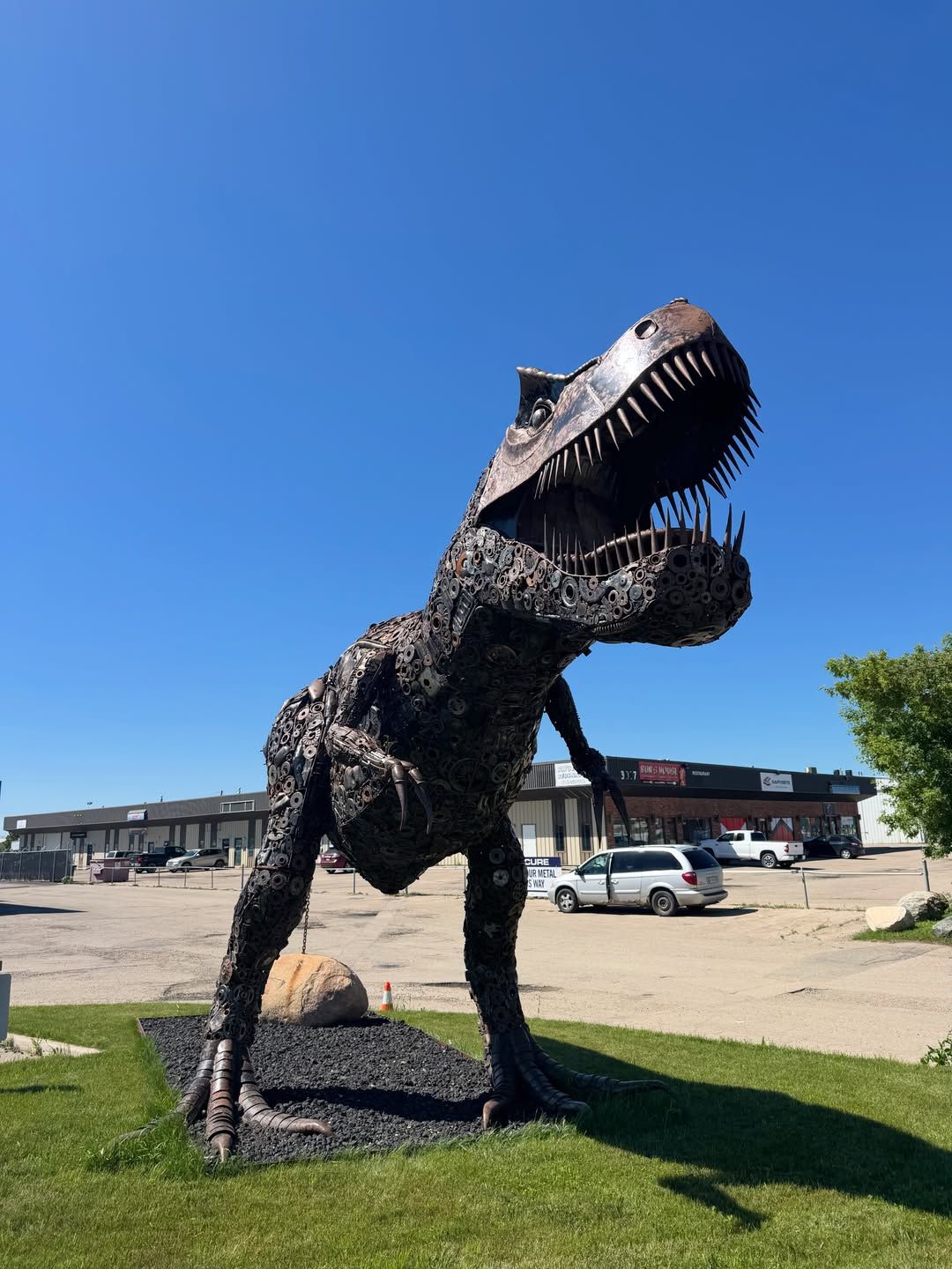 Large metal dinosaur sculpture with open mouth in a parking lot.