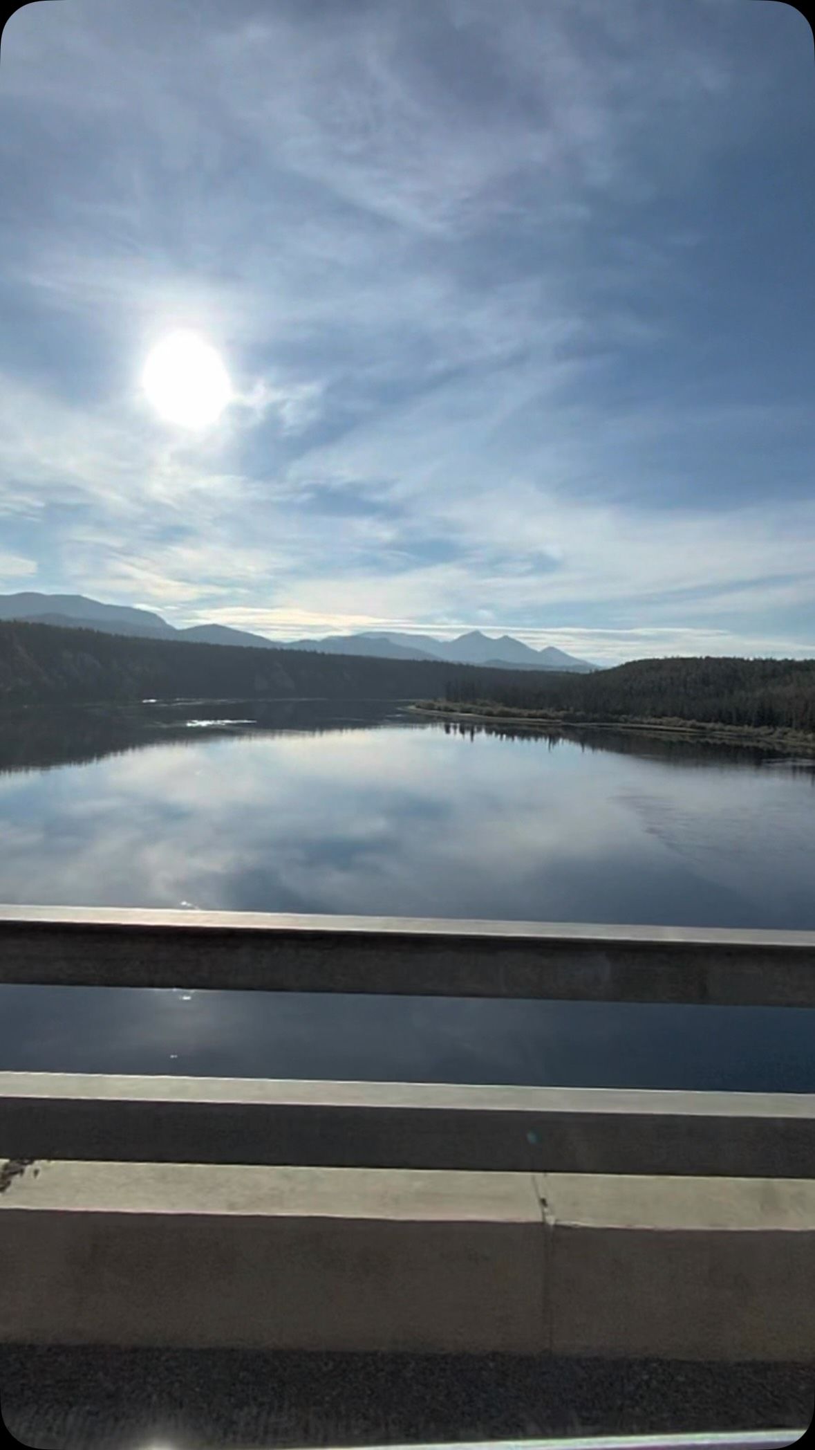 Still lake reflects mountains and forest under blue sky from van window.