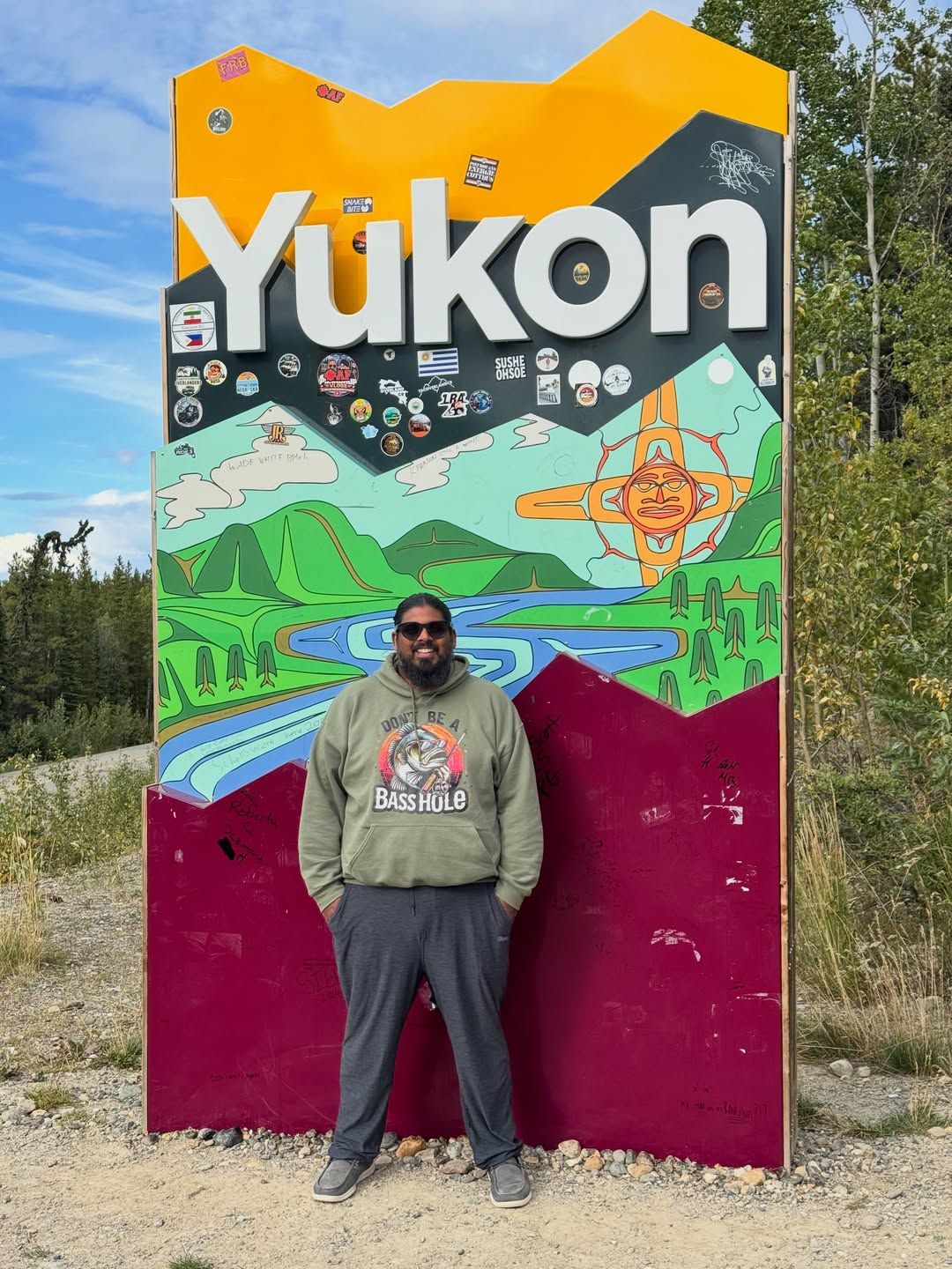 Man in hoodie and sunglasses standing in front of colorful Yukon welcome sign.