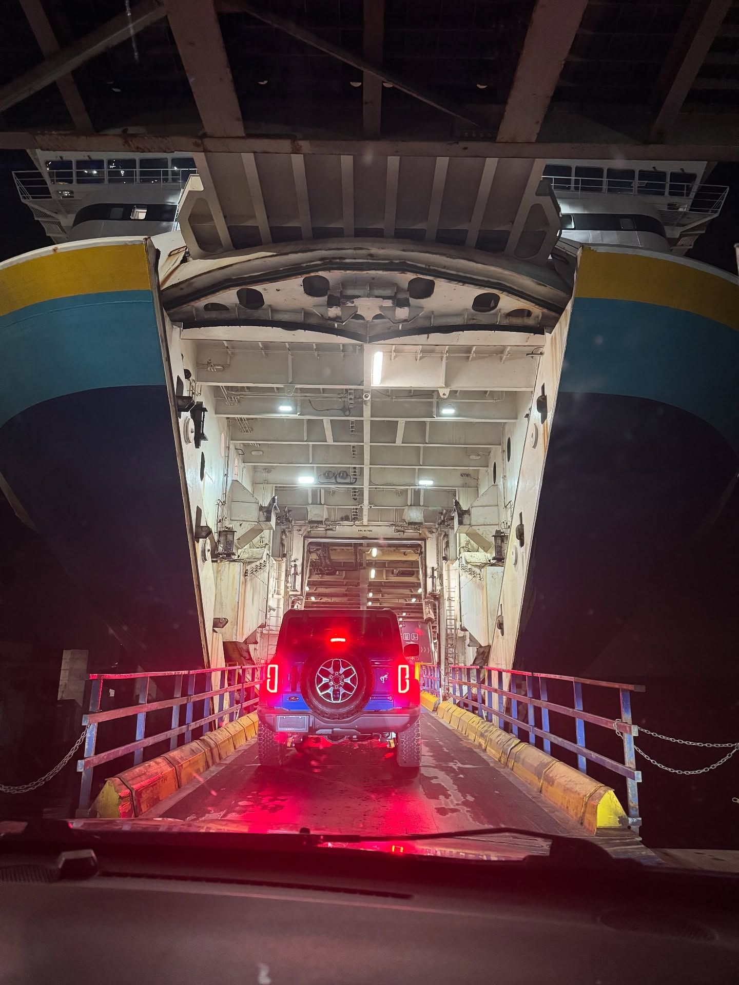 Vehicle deck of ferry with illuminated red Jeep and metal railings at night.