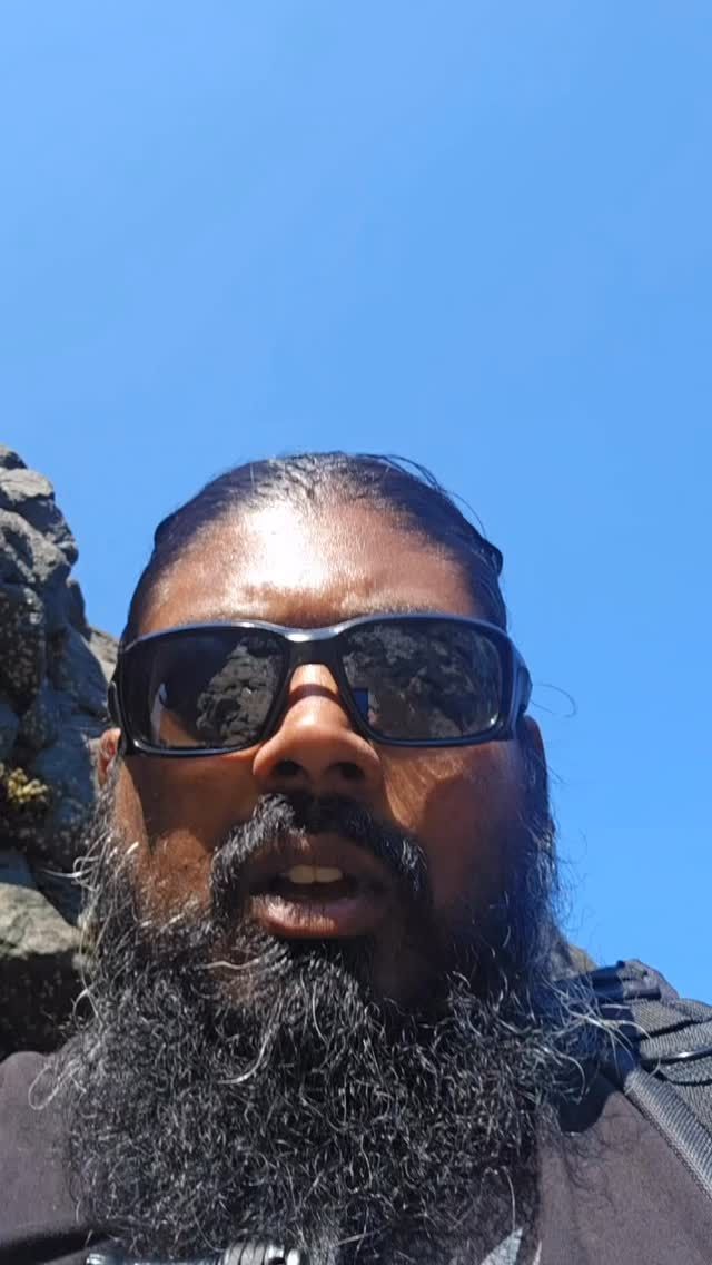 Man with beard and sunglasses smiles for selfie against blue sky and rocks.