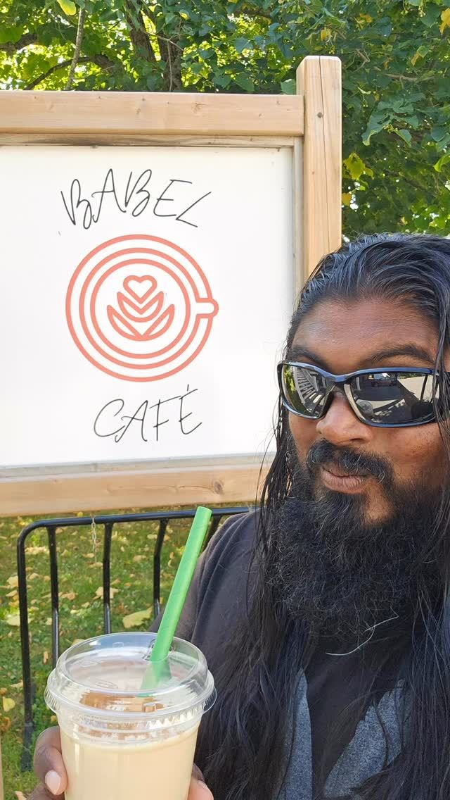 Man holding iced drink at outdoor cafe with wooden sign visible.