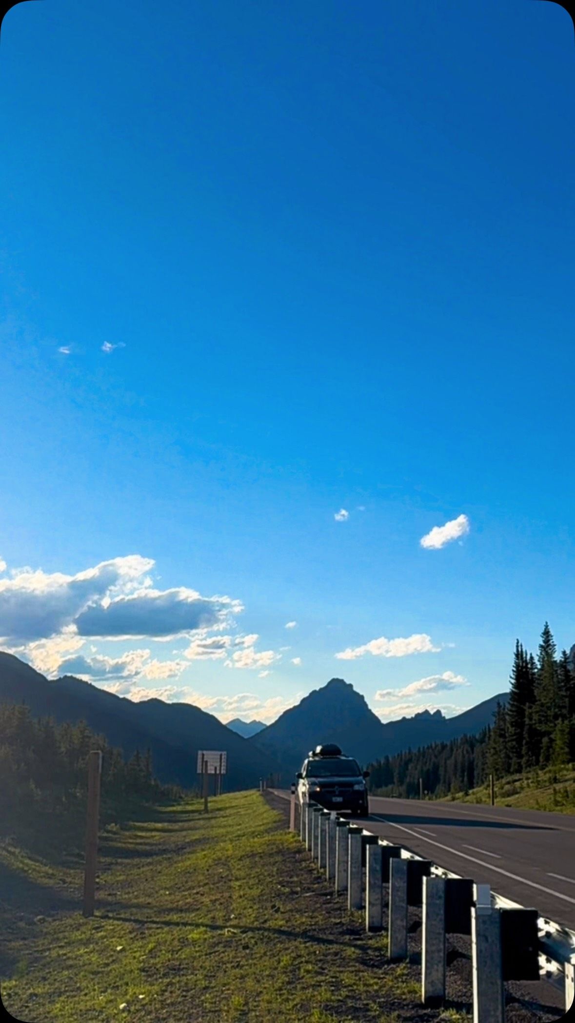 Van driving on mountain highway through evergreens under bright blue sky.
