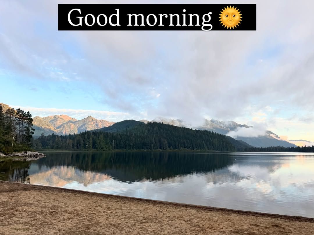 Mountain lake with forested shoreline reflects dramatic sky at sunrise.