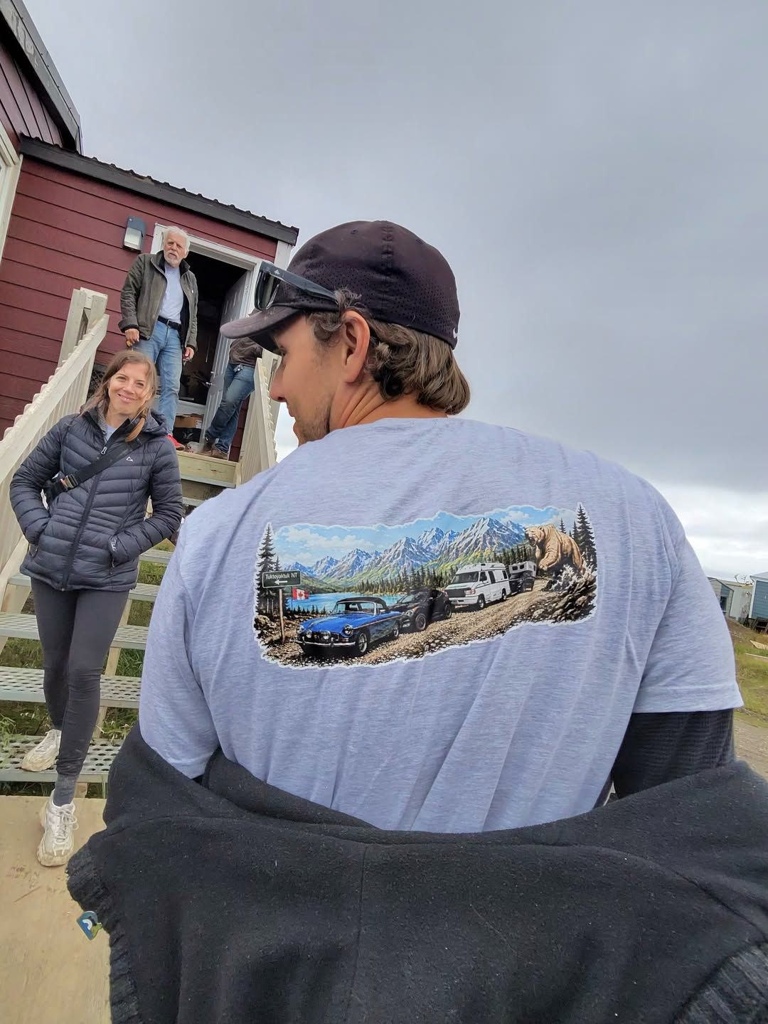 Two people stand outside a red cabin, one wearing a vintage van graphic shirt.