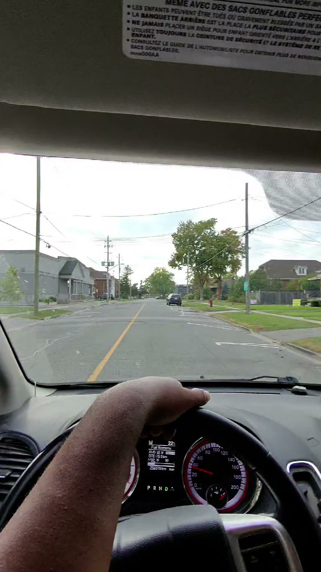 Driver's hand on wheel on residential street lined with houses and leafy trees.