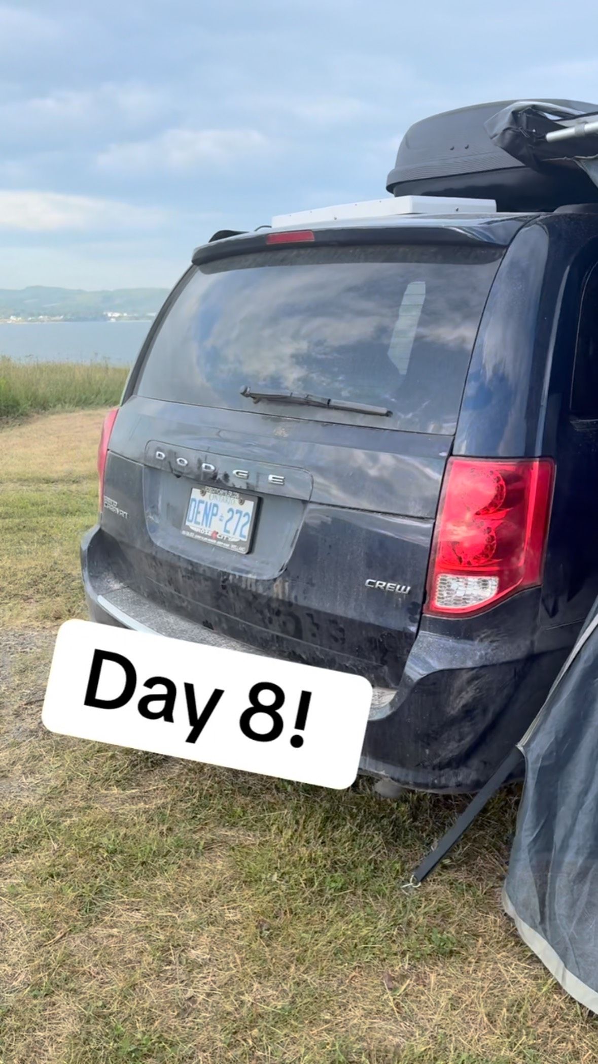 A parked minivan sits by a grassy shoreline with water and distant hills.
