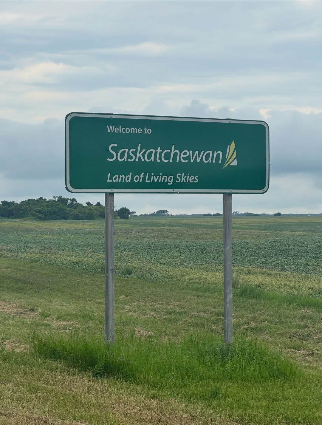 Roadside sign welcomes travelers to Saskatchewan with prairie landscape behind.