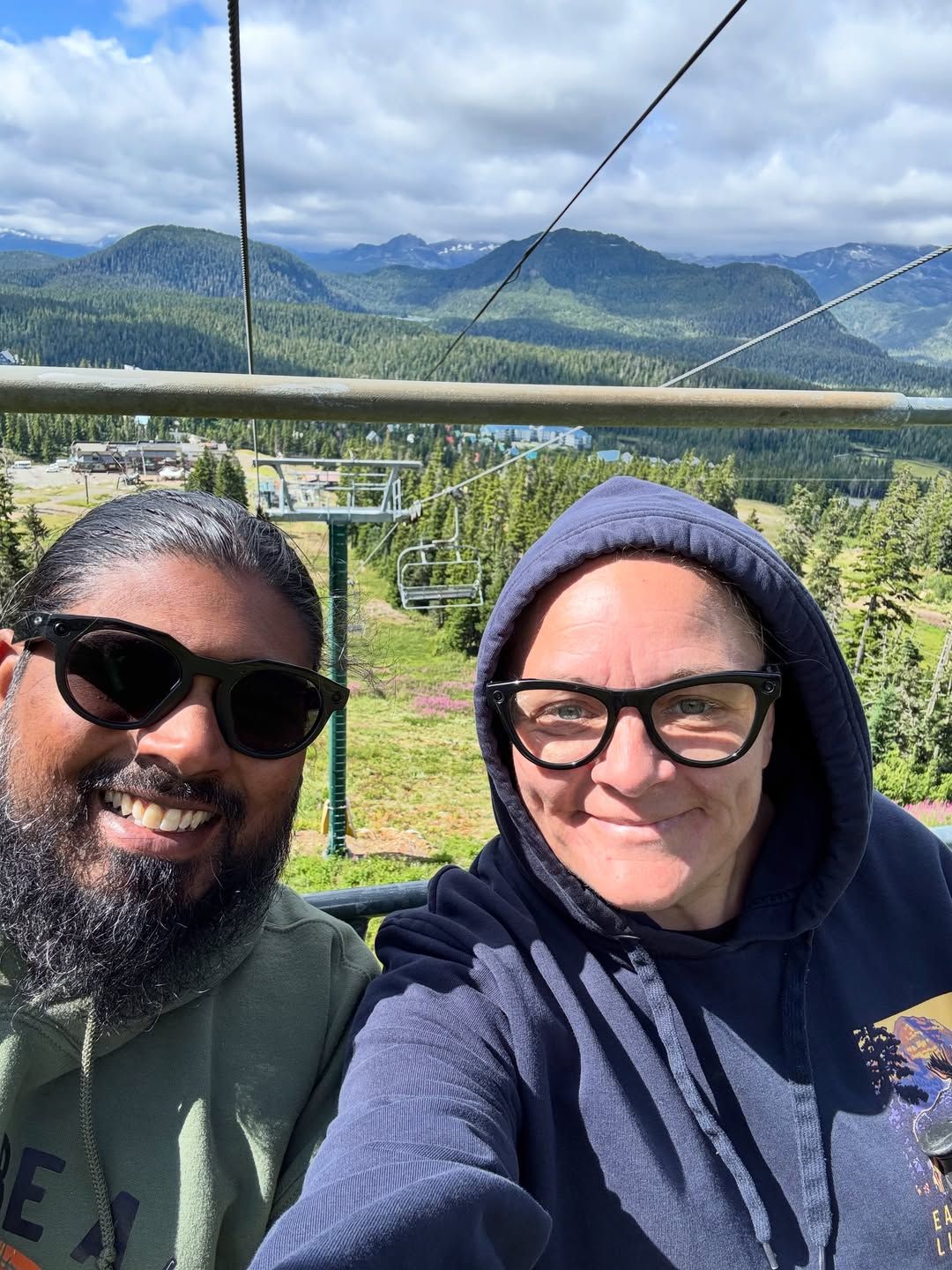 Two people on ski lift with forested mountain valleys below.